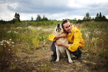 Arka planda bulutlu bir gökyüzü ve çiçek açan çayırda bej renkli Labrador av köpeğiyle oturan genç adamın yan görüntüsü.