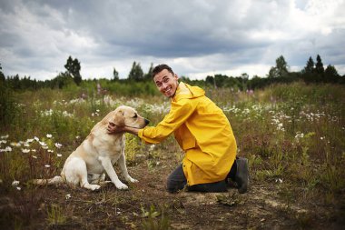 Labrador Retriever 'ın önünde tek dizinin üstünde dururken gülümseyerek gülen rahat genç bir adamın yan görüntüsü