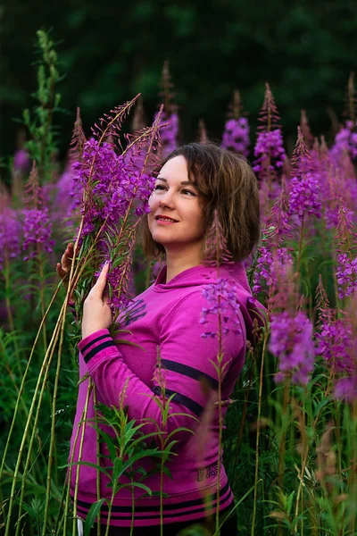 Plus size woman in fuchsia hoodie looking aside while standing amidst bright flowers in meadow in countryside