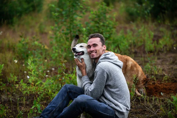 stock image Side view of happy man in casual clothes looking at camera and hugging dog while sitting on ground during walk in forest