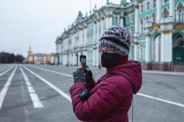 Siyah maskeli genç bir kadının akıllı telefondan ateş edişinin yan görüntüsü. Kentin merkezindeki boş caddelerde kendini izole ediyor.