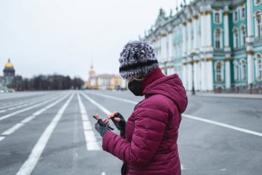 Siyah maskeli genç bir kadının akıllı telefon yazışının yan görüntüsü. Şehrin merkezindeki boş sokaklar arka planda kendini izole ediyor.