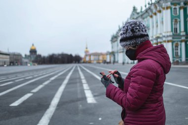 Siyah maskeli genç bir kadının akıllı telefon yazışının yan görüntüsü. Şehrin merkezindeki boş sokaklar arka planda kendini izole ediyor.