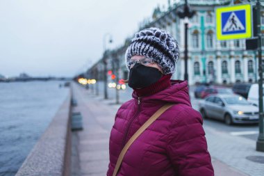 Siyah maskeli genç bir kadın setin kenarında dikilip yan bakıyor. Şehrin boş caddesi, arka planda karantina altında