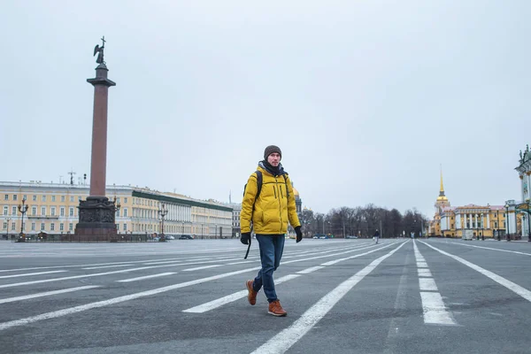 Side view of Young man in yellow jacket walking on the road. Empty dvortsovaya square in the centre of the city on background