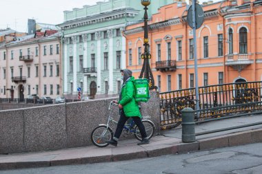 Saint-petersburg, Rusya - 31 Mart 2020: Yeşil ceketli ve maskeli kurye, şehirde bisikletle siparişleri teslim ediyor.