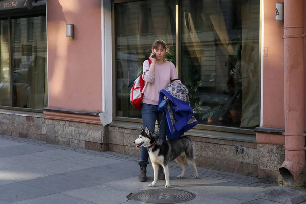 2020年4月7日 俄罗斯圣彼得斯堡 一位年轻貌美的女士带着一只哈士奇犬站在人行道上 与人通电话 — 图库照片