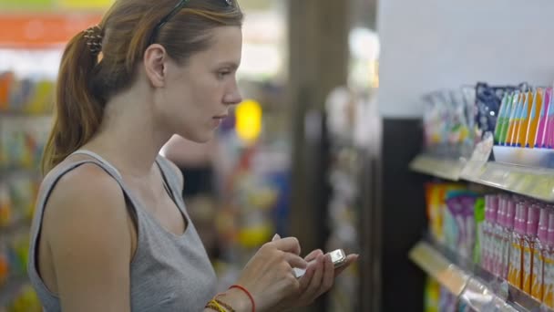インドネシアのバリ- 2019年3月:化粧品店でスマートフォン読み取り製品ラベルを使用している若いアメリカ人女性. — ストック動画
