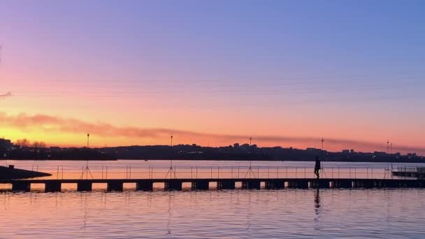 日の出、日没時に湖の上に夕暮れの空と橋を渡って歩く人のシルエットビュー. — ストック動画