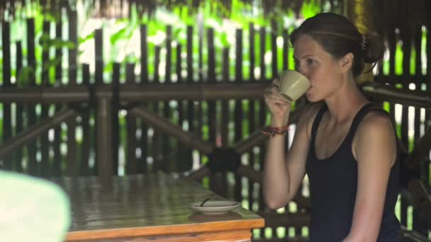 Ung kaukasisk kvinna dricker kaffe vid bordet på terrassen på den tropiska ön. — Stockvideo