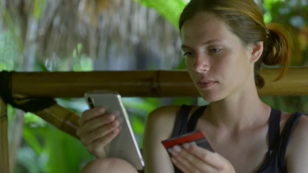 Mulher segurando cartão de crédito e usando telefone celular. feminino compras on-line em resort exótico ao ar livre . — Vídeo de Stock