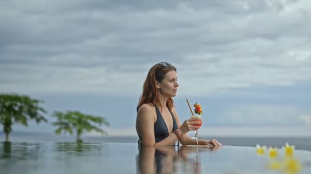 Pretty young woman drinking cocktail in edgeless pool on resort hotel. female tourist swimming — Stock Video