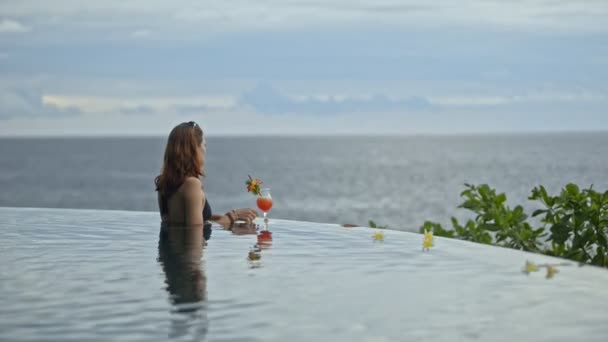 Ung rödhårig kvinna dricker cocktail och tittar på havet landskap i poolen på Bali Island. — Stockvideo