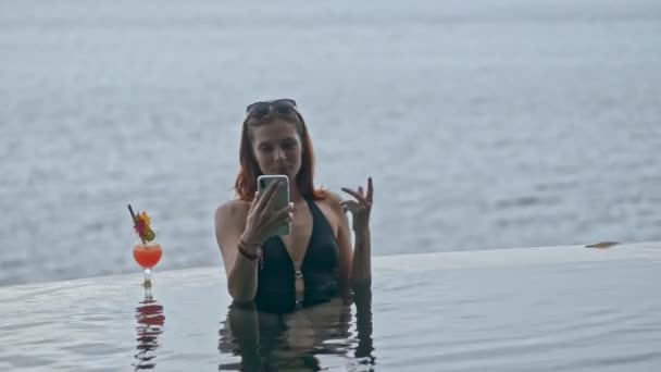 Beautiful red haired woman having video call using smartphone in ocean pool at tropical resort. — Stock Video