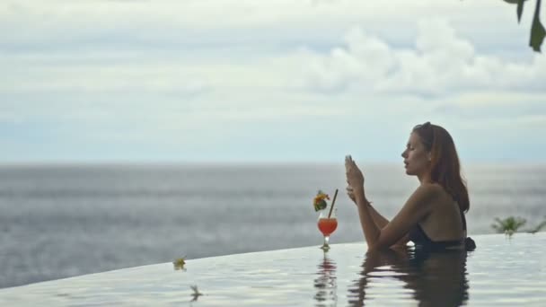 Вид сбоку на сексуальную женщину, пьющую коктейль в бассейне бесконечности в отпуске. разговор через facetime видео-звонок — стоковое видео