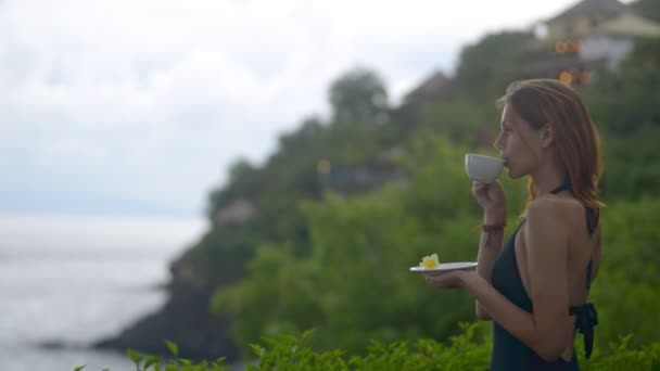 Frau im bali hotel resort trinkt Tee oder Kaffee und blickt auf das Meer vor dem regnerischsten Hintergrund. Morgen — Stockvideo