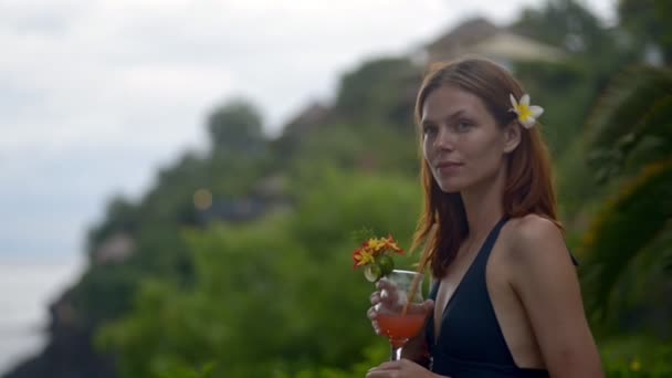 Schöne junge rothaarige Frau posiert mit einem Cocktail in der Hand, während sie auf einer tropischen Insel steht. — Stockvideo