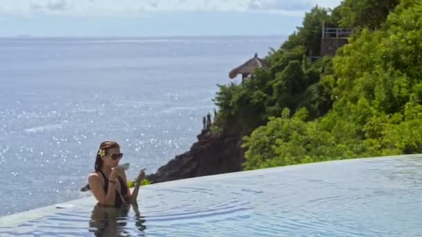 Woman in infinity pool drinking coffee and holding mobile phone. pretty sexy female relax — Stock Video