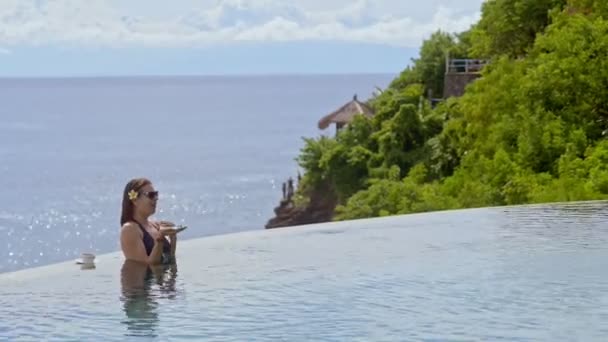 Joven mujer caucásica hablando sosteniendo teléfono inteligente en la mano, descansando en la piscina en la isla . — Vídeos de Stock