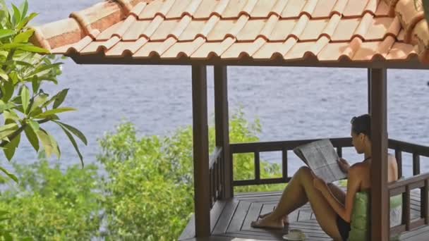 Junge kaukasische rothaarige Frau, die Zeitung liest, sitzt auf einer Terrasse auf einer tropischen Insel. — Stockvideo