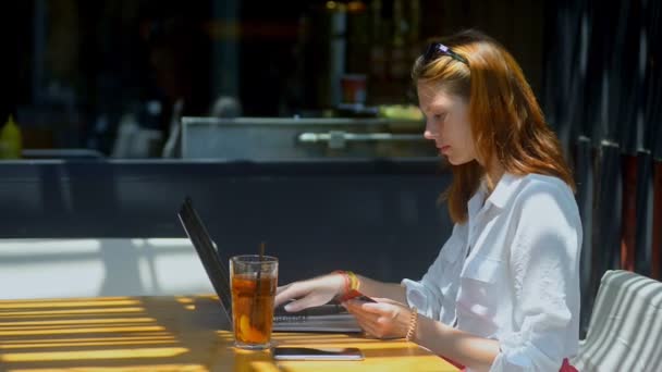 Молодая рыжая женщина с помощью ноутбука и карточки сидя в летнем кафе во время путешествия . — стоковое видео