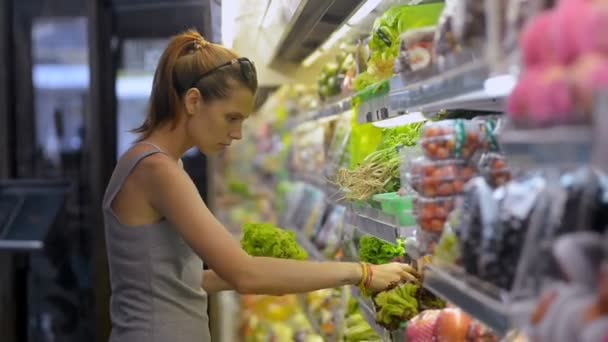 Jonge roodharige vrouw kiezen en kopen bladgroenten tijdens het winkelen in biologische winkel. — Stockvideo