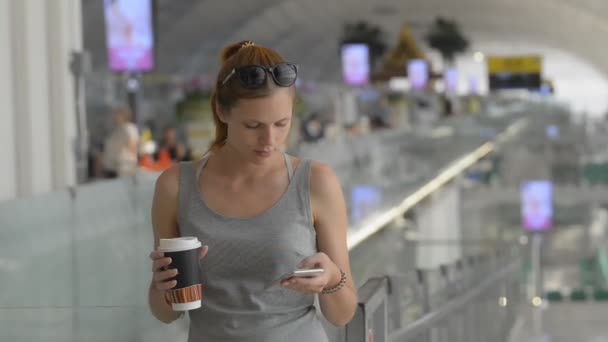 Young red haired woman using smartphone and holding coffee in hand standing in modern mall. — Stock Video