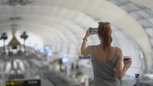 Vista posterior mujer caucásica viajero tomando fotos en el aeropuerto. celebración de teléfono móvil en la terminal de espera — Vídeos de Stock