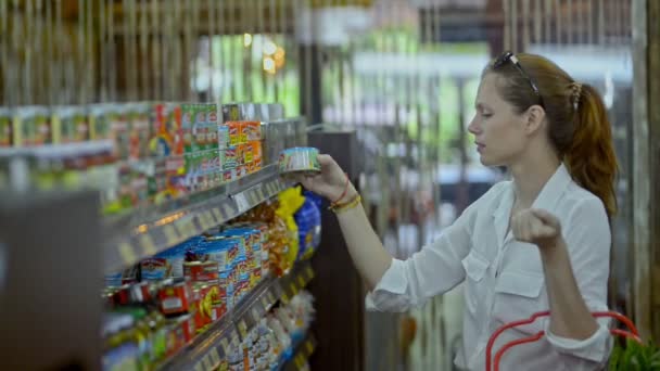 Bali, Indonesia - Marzo, 2019: Joven mujer pelirroja caucásica eligiendo comida enlatada de pie en el supermercado moderno durante un viaje asiático . — Vídeo de stock