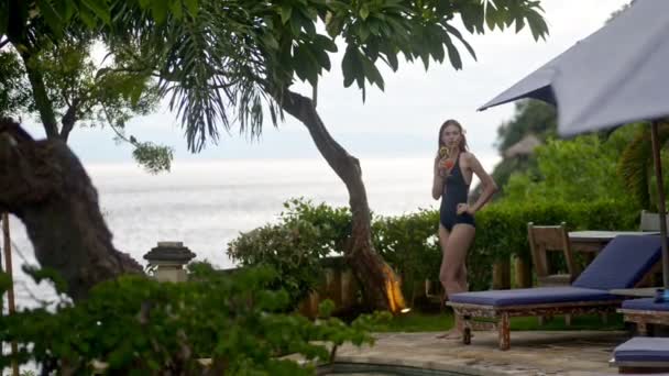 Gyönyörű nő modell iszik egzotikus koktél, miközben áll a tengerparton a trópusi szigeten. — Stock videók