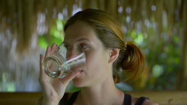 Porträt einer jungen kaukasischen rothaarigen Frau, die Wasser trinkt, sitzt in einem tropischen Café im Freien. — Stockvideo
