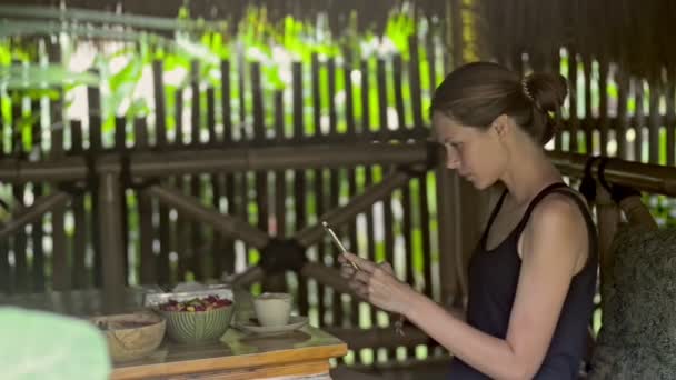 Jonge roodharige vrouw met smartphone en het drinken van thee aan tafel in eiland cafe. — Stockvideo