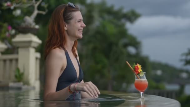 Mujer joven en una piscina infinita bebiendo cóctel. nice female relax laugh smile on rainforest background. — Vídeo de stock