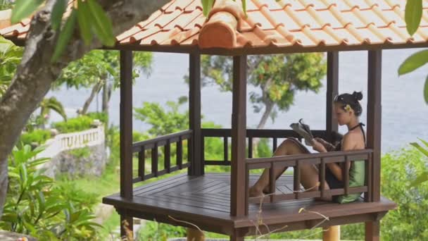 Young red haired woman reading newspaper and drinking tea sitting at terrace of asian resort. — Stock Video