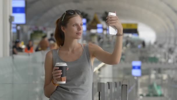 Giovane viaggiatore caucasico donna che prende selfie in aeroporto. in possesso di telefono cellulare e tazza di carta da caffè — Video Stock