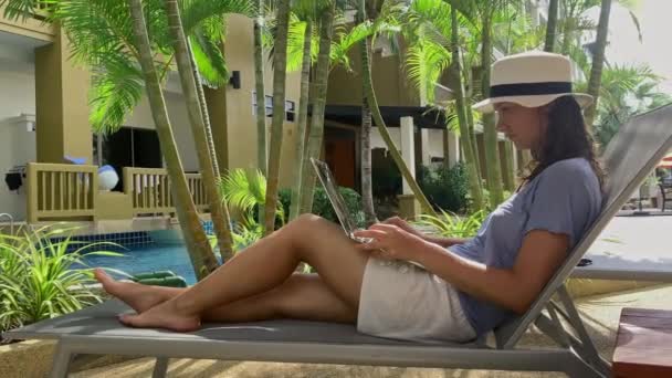 Фрілансерка посміхається і працює на ноутбуці біля басейну. бізнес-леді у відпустці — стокове відео