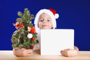 Genç Beyaz kutu arkasından bir hediye arıyorsunuz Noel Baba. Hugs hediye ve bir Noel ağacı. Kameraya bakıyor. Mavi arka plan. Noel ağacı masanın üzerinde duruyor. Chromakey. Yakın çekim.