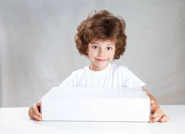 Pequeño niño lindo rizado con una caja blanca en sus manos mirando tiernamente a la cámara. Fondo gris. Primer plano . — Foto de Stock