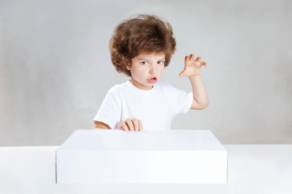 Pequeño chico de pelo rizado lanza un hechizo sobre la caja blanca. Fondo gris. Primer plano . — Foto de Stock