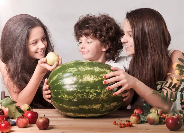 Three children playing with fruits sitting at the table in front of a huge watermelon. Gray background. Close-up. — Stock Photo, Image