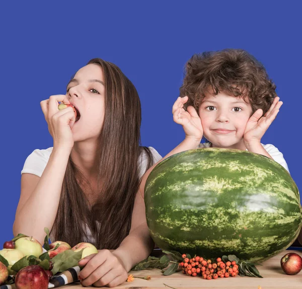 Brother and sister sitting at a table. Wait a delicious watermelon. Sister eating apple. Brother smiling and looking at the camera. Blue background. Close-up. — Stock Photo, Image