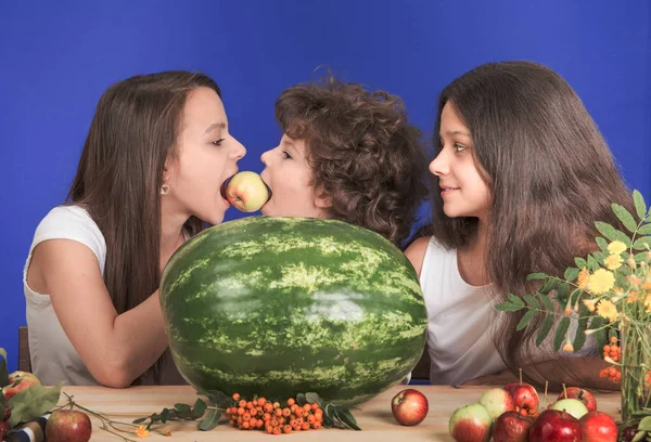 Két nővére és a little brother ül az asztalnál. Az asztalon van egy hatalmas egész görögdinnye és a gyümölcs. Testvérpár csípős egy alma. Közeli kép:. — Stock Fotó