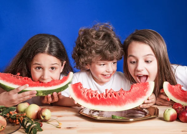 Little curly boy and two girls sitting at a wooden table and eat large ripe watermelon. Blue background. Close-up. — Stock Photo, Image