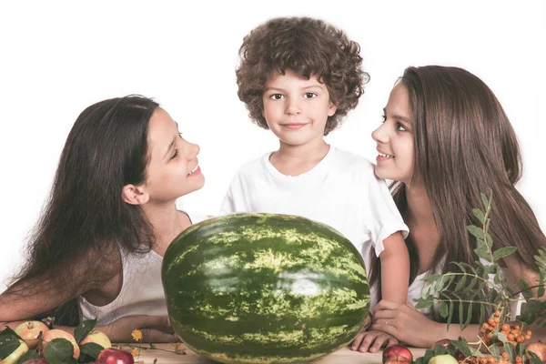 Három gyermek ül az asztal egy nagy görögdinnye egész. A fiú úgy néz ki, a kamera. A lányok egymásra nézni. Fehér háttér. Közeli kép:. — Stock Fotó