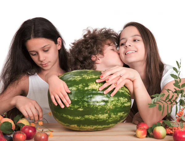 Három gyermek ül az asztalnál. A fiú nagy görögdinnye tegye a kezét, és megcsókolja a lányt, az arcát. Fehér háttér. Közeli kép:. — Stock Fotó
