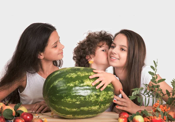 Három gyermek ül az asztalnál. A fiú kezét egy nagy görögdinnye és suttogja a fülébe egy lány. Szürke háttér. Közeli kép:. — Stock Fotó