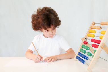 Sevimli kıvırcık saçlı çocuk kurşun kalemle kağıt üzerinde yazıyor ve sonuçlarına görünüyor. Yakın çekim. Gri arka planı.