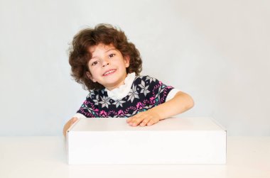 Mutlu sevimli çocuk kameraya bakarak bir gülümsemeyle Başını eğdi. Beyaz kutu bir yandan saklıyor. Gri arka planı.