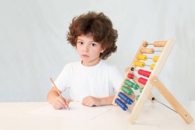 Üzgün şirin küçük çocuk dalgın suratında bir kağıt bir kalem yazar. Kameraya bakıyor. Masaya oturur. Gri arka planı.