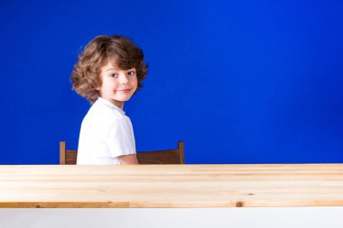 Kafasını çevirerek yarı açık, gülümseyen ve seyir kameranın içine oturan mutlu kıvırcık şirin çocuk. Yakın çekim. Mavi arka plan.